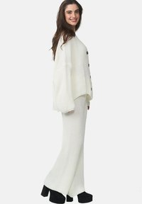 Ensemble en maille blanche composé d'un pull à rayures et d'un pantalon évasé avec des boutons sur le devant, offrant une coupe décontractée et un tissu texturé. Des chaussures à plateforme noires complètent le look.
