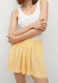 Shorts floraux jaunes avec un waistband élastique, fabriqués en tissu léger, ornés de petits motifs de fleurs blanches et violettes.