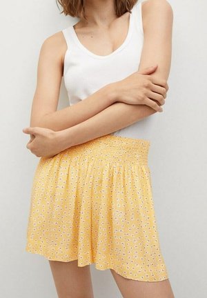Shorts floraux jaunes avec un waistband élastique, fabriqués en tissu léger, ornés de petits motifs de fleurs blanches et violettes.