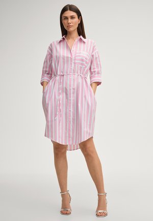 Femme portant une robe chemise à rayures verticales roses et blanches avec des poches et des sandales argentées à talons hauts, debout les mains dans les poches.