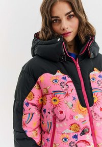 Podšívková bunda s čiernou hornou časťou a živou ružovou dolnou časťou, pokrytou rôznymi nebeskými motívmi, vrátane slnka, mesiacov a hviezd. Zips a kapucňa.
