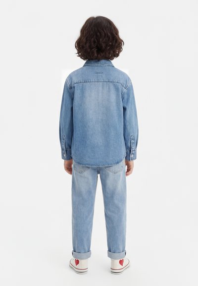 Enfant aux cheveux bouclés et foncés, debout de dos, portant une veste en jean bleu clair et un jean, chaussures blanches avec un détail cœur rouge.