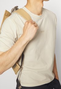 Světle béžové bavlněné tričko s kulatým výstřihem, na kterém je jemné logo "BOSS". Na jednom rameni je viditelný hnědý popruh od batohu.