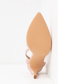 Chaussure à talon haut en cuir nude avec un bout pointu, des lanières fines à la cheville et une semelle en caoutchouc texturée, présentant une finition lisse et un design effilé.