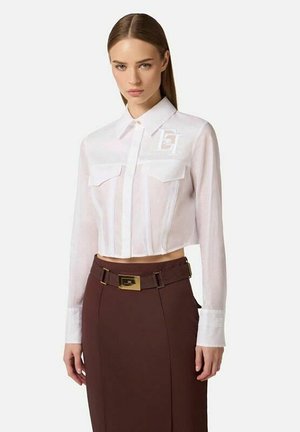 Donna che indossa una camicia bianca corta con bottoni e tasche sul petto, abbinata a una gonna marrone a vita alta con una cintura larga e fibbia dorata.