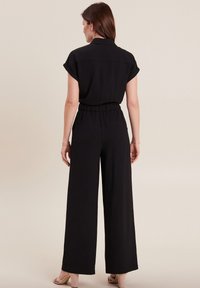 Zwarte broekjumpsuit met brede pijpen, korte opgerolde mouwen, elastische tailleband en achterzakken. Gladde textuur en een minimalistisch ontwerp.