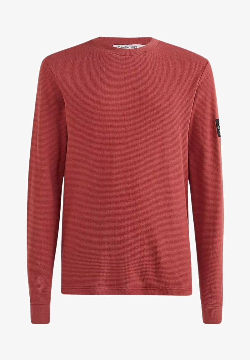 Pull rouge à manches longues avec un tricot texturé, col rond et écusson noir sur la manche gauche. Coupe standard, poignets et ourlet côtelés.
