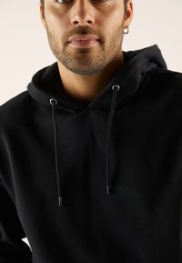 Schwarzer Hoodie aus weichem Stoff, mit einem Kordelzug an der Kapuze, silbernen Ösen und einer lockeren Passform. Nahaufnahme mit Fokus auf den Halsausschnitt.