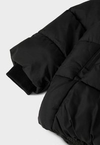 La manica della giacca a vento nera presenta una texture trapuntata, un polsino elasticizzato e una finitura in tessuto morbido con un dettaglio della tasca con zip.