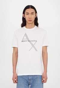 Fehér pamut póló, grafikával, melyen a "ARMANI EXCHANGE" felirat és stilizált "A" és "X" sötétszürke színben található, rövid ujjal és kerek nyakkivágással.