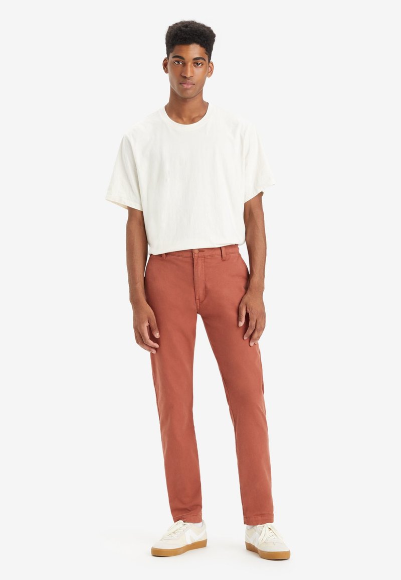 T-shirt blanc à manches courtes avec une coupe décontractée, associé à un pantalon droit couleur rouille. Le modèle porte des baskets blanches avec des accents beiges.