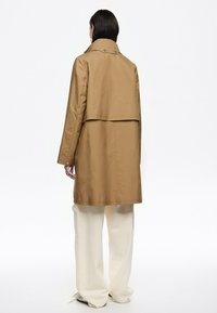 Cappotto lungo marrone con uno spacco posteriore e dettagli con bottoni. Il cappotto ha una superficie liscia ed è abbinato a pantaloni larghi color crema.