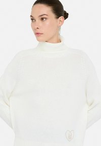 Maglione bianco a collo alto con texture a costine e vestibilità oversize. Presenta un discreto logo a forma di cuore sul lato inferiore. Tessuto a maglia liscia.