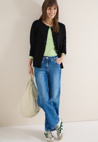 Femme debout contre un mur uni, portant un cardigan noir, un haut vert clair, un jean bleu, des baskets blanches et vertes, tenant un sac beige.