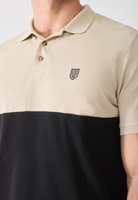 Beige og svart polo skjorte med to-farget design, teksturert stoff, tre-knaps placket og en liten brodert logo.
