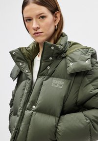 Veste matelassée verte avec un col montant, dotée d'une fermeture éclair, de boutons-pression et d'une étiquette de marque discrète sur la poitrine. Texture lisse.