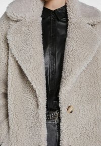 Gros plan sur un manteau sherpa beige porté sur une chemise en cuir noire à boutons, une ceinture noire cloutée et un jean en denim gris.