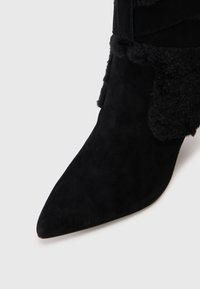 Stivale in suede nero a punta con un morbido bordo in pelliccia lungo la caviglia su uno sfondo bianco.