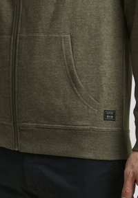 Oliven grønn zip-up hettegenser laget av teksturert stoff, med kengurulomme og ribbestrikkede ermer, med et lite merke på siden.