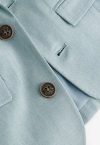 Blazer azzurro chiaro con finitura testurizzata, dotato di due tasche e un bottone marrone con testo inciso. Fodera interna di colore azzurro chiaro.
