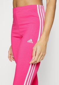 Rosa träningstights i stretchigt material, med tre vita ränder längs sidan och en Adidas-logotyp vid höften.