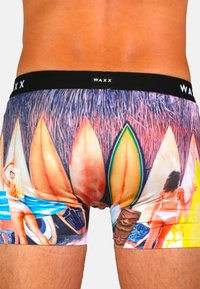 Shorts de bain avec un imprimé vif représentant une scène de surf et de plage, fabriqués en tissu extensible, avec une ceinture noire WAXX et des graphiques colorés.