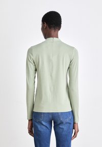 Langermet topp i lys grønn stoff, med glidelås bak, glatt tekstur og en tettsittende silhuett, kombinert med blå jeans.