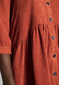 Robe en velours côtelé couleur rouille avec ouverture à boutons sur le devant, taille froncée et manches courtes. Comprend des boutons bleus contrastants et une texture côtelée verticale.