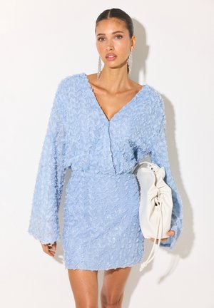 Femme portant une robe texturée bleu clair, tenant un sac à main blanc plissé, avec de longues boucles d’oreilles et les cheveux tirés en arrière, debout devant un fond uni.