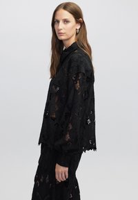Blusa nera in pizzo con maniche lunghe, colletto alto e motivi floreali con sezioni trasparenti. Il tessuto è texturizzato con dettagli intricati.