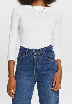 Frau trägt ein weißes, eng anliegendes Langarmshirt, hoch taillierte blaue Jeans mit doppeltem Knopfverschluss, goldene Halskette und goldenes Armband.