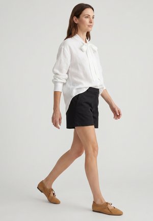 Frau geht und trägt eine weiße Langarmbluse mit Schleife, schwarze Shorts und beige flache Schnürschuhe vor einem schlichten hellen Hintergrund.