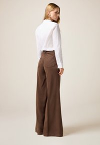Camisa blanca de botonadura frontal combinada con pantalones anchos marrones, que cuentan con bolsillos traseros y una textura suave. El modelo se presenta con un fondo neutro.