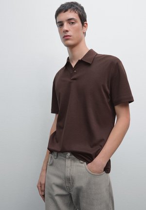 Brun polo shirt med klassisk krave, korte ærmer og to knapper. Båret med lysegrå jeans, sat mod en neutral baggrund.