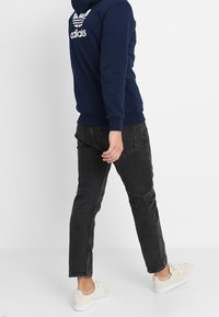 Marinblå hoodie med vitt Adidas-logotyp på bröstet, kombinerad med svarta jeans och ljusa sneakers, sedd bakifrån mot en enfärgad bakgrund.