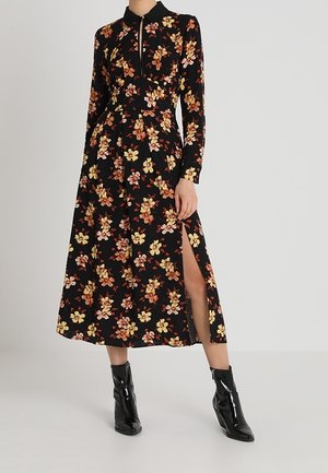 Femme portant une robe noire à manches longues avec un imprimé floral orange et jaune, fendue sur le devant, associée à des bottines noires en cuir verni.