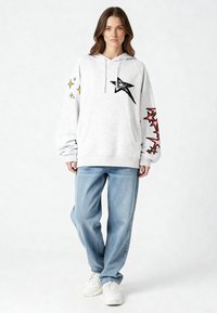 Felpa con cappuccio grigio chiaro con tasca frontale, caratterizzata da una grafica di una stella nera e accenti di stelle colorate sulle maniche. Abbinata a jeans azzurri e sneakers bianche.