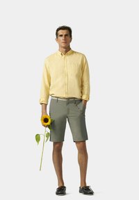 Chemise beige clair à boutons, short vert, chaussures noires. Tient un tournesol, avec des détails décoratifs sur la poche et la ceinture du short.