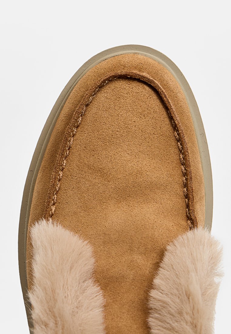 Mocassin en suède beige avec un bout arrondi, arborant un accent en fourrure beige duveteuse le long du bord et une semelle en caoutchouc de couleur claire.