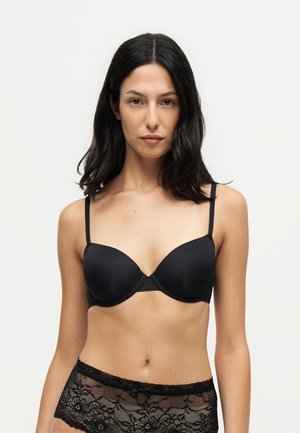 Femme aux cheveux foncés mi-longs portant un soutien-gorge noir et une lingerie noire en dentelle sur un fond clair uni.