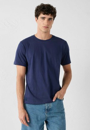 Navyblauwe katoenen T-shirt met een klassieke ronde hals, korte mouwen en een subtiele logo-detail op de zoom, gecombineerd met lichtblauwe jeans.