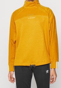 Keltainen fleece-pullover korkealla kauluksella, jossa edessä vetoketju ja logo, musta kiristysnauha helmassa sekä raidalliset yksityiskohdat.