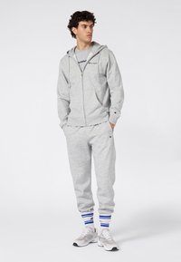 Sudadera gris con capucha y cremallera frontal, combinada con pantalones deportivos grises a juego. Incluye calcetines tobilleros con rayas azules y zapatillas deportivas grises.