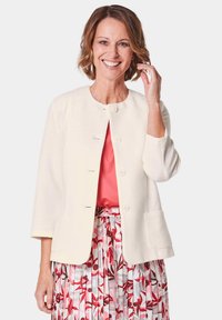 Witte textuurjacket met ronde hals, zes knopen en twee voorzakken, gecombineerd met een roze blouse en een gestreepte, geplooide rok.