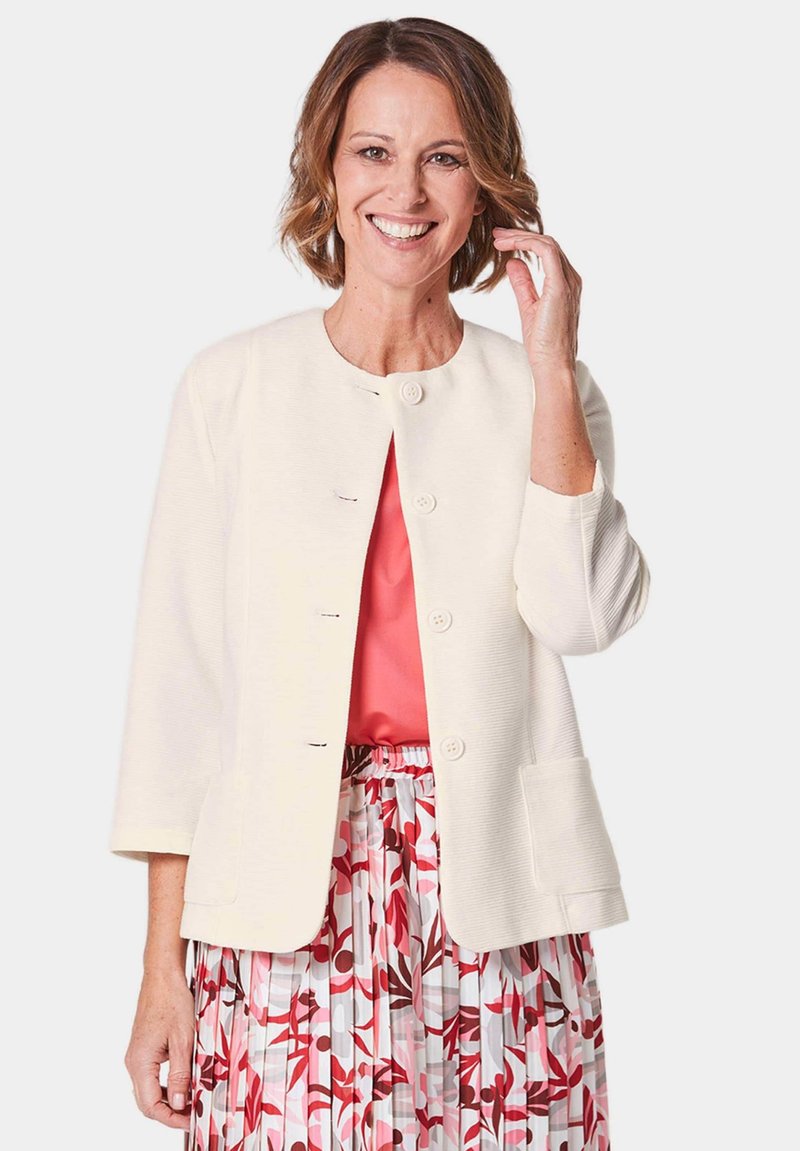 Weiß strukturierte Jacke mit rundem Kragen, sechs Knöpfen und zwei Vorder- taschen, kombiniert mit einer rosa Bluse und einem gemusterten, plissierten Rock.