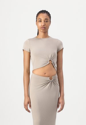 Kobieta z warkoczami, ubrana w beżowy krótki crop top z krótkim rękawem i pasującą spódnicę, obie z okrągłymi wycięciami w kształcie pierścienia na wysokości talii.