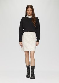 Schwarzer Pullover mit geprägtem Text, kombiniert mit einem strukturierten weißen Rock. Das Model trägt schwarze Knöchelstiefel mit grauen Socken und steht vor einem hellen Hintergrund.