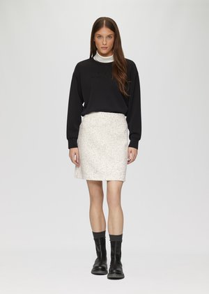 Sweatshirt noir avec texte en relief, associé à une jupe blanche texturée. Le modèle porte des bottines noires avec des chaussettes grises, se tenant devant un fond clair.