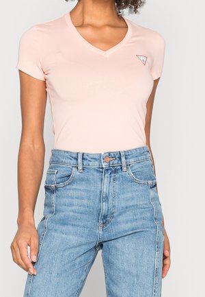 Kvinde iført lyserød V-udskåret T-shirt med korte ærmer og lille logo på brystet samt højtskårede lyseblå jeans mod en ensfarvet baggrund.