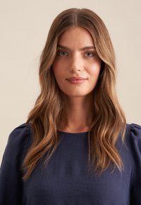 Navybluse mit rundem Halsauschnitt, gerafften Puffärmeln und einem strukturierten Stoff. Lange, wellige braune Haare umrahmen das Gesicht. Neutraler Hintergrund.